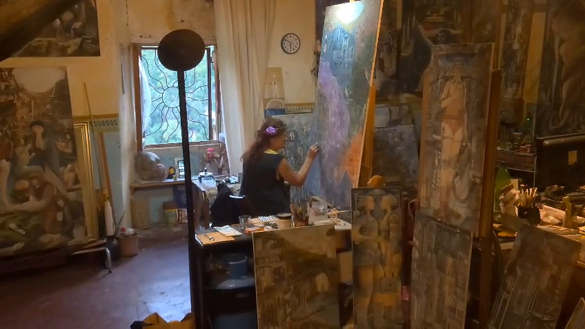 Angela dipinge al cavalletto, piccola all’interno dello studio pieno di tele, oggetti e luce dalla finestra.